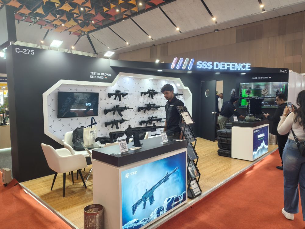 The SSS Defence stall on the show floor: a black booth with a lit SSS Defence fascia, a white hexagon-patterned wall of rifles, and T12 and Varaha display counters
