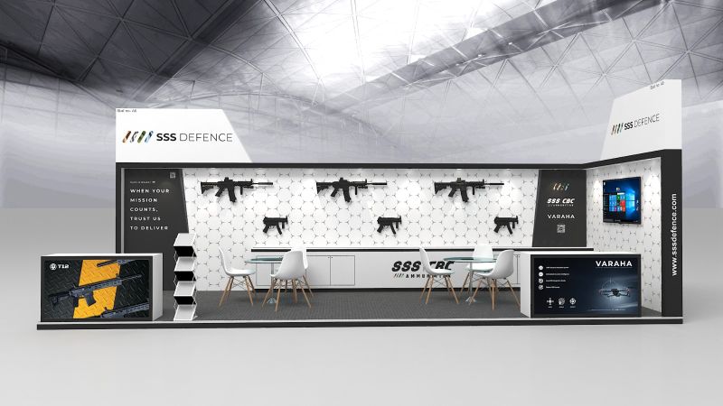 Render of an open corner stall: white hexagon wall with rifles and submachine guns, a black panel reading When your mission counts, trust us to deliver, and T12 and Varaha counters